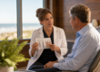 A female dentist explains a treatment protocol to a male patient seated in a dental chair near a sunlit window.