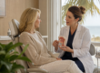 A dentist sits beside a patient near a sunlit window, gently explaining an airway model during a relaxed consultation.