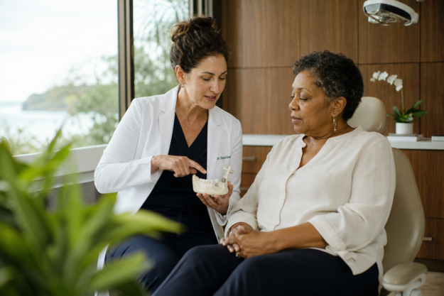 A dentist in her 40s explains jaw anatomy to an attentive patient using a small model in a warmly lit dental room.